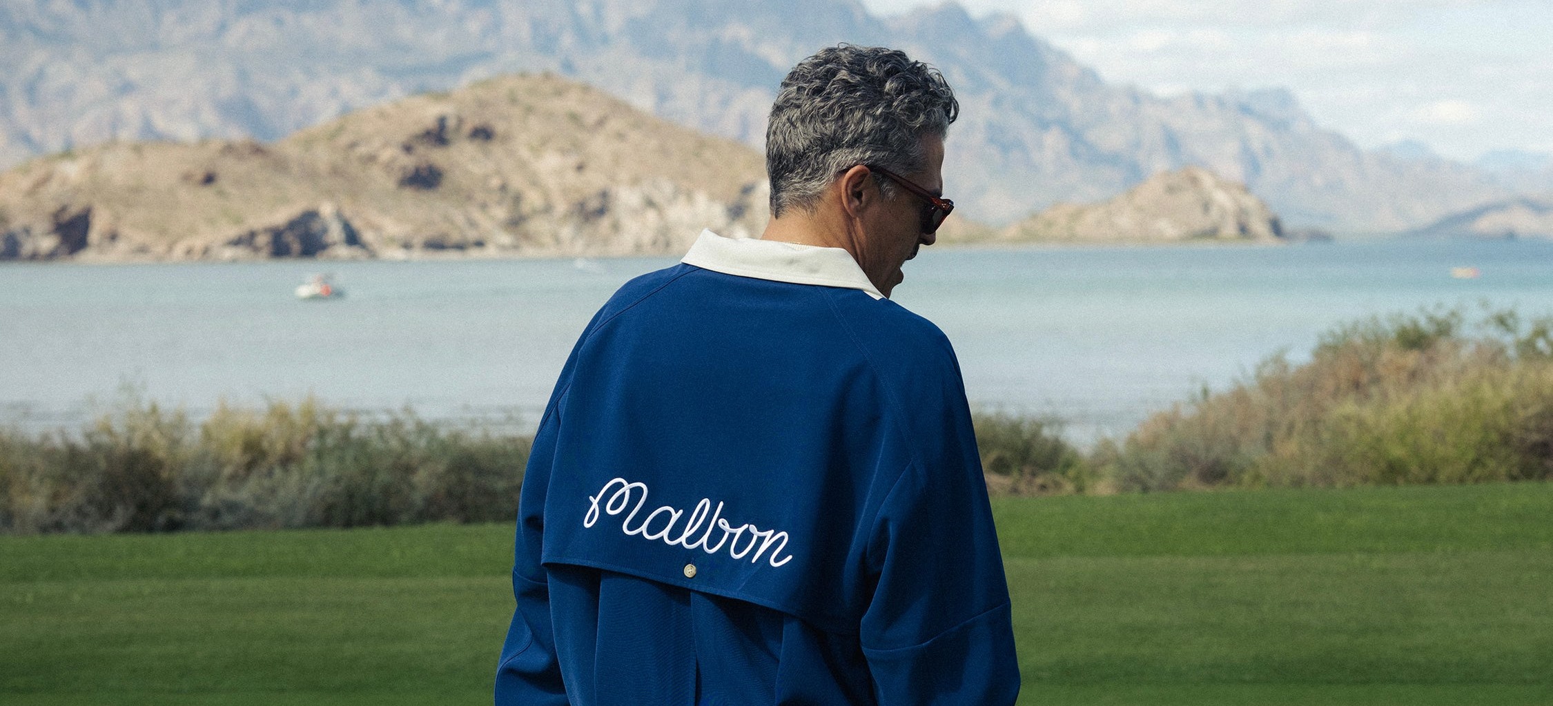 Malbon 創辦人形象:藍色外套背後的白色 Malbon 草寫刺繡,身後是海灣球場