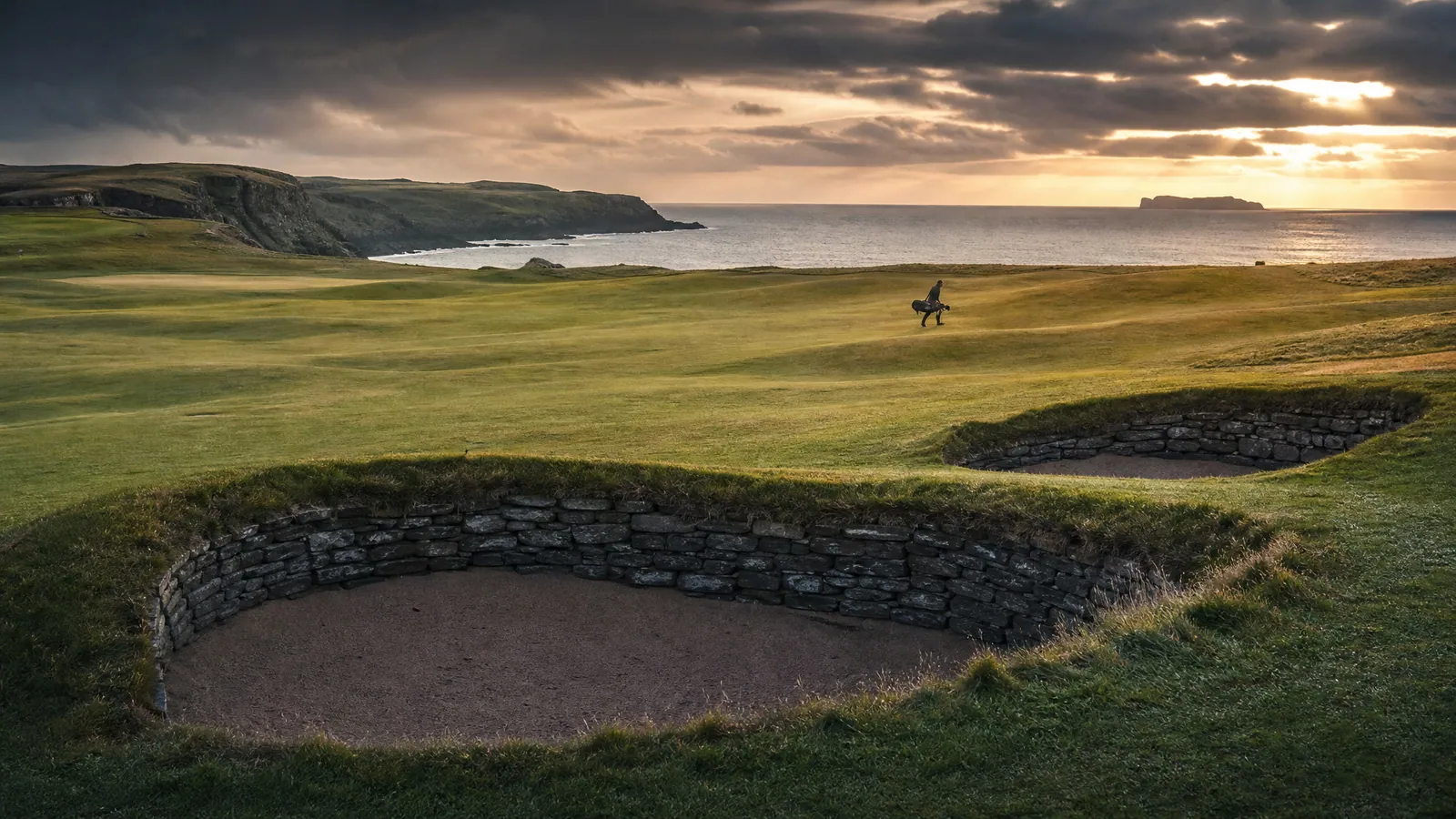 蘇格蘭 links 球場的傳統石頭 pot bunker 兩個，遠處球員揹球袋走過 fairway，背景是海岸線與戲劇性夕陽雲