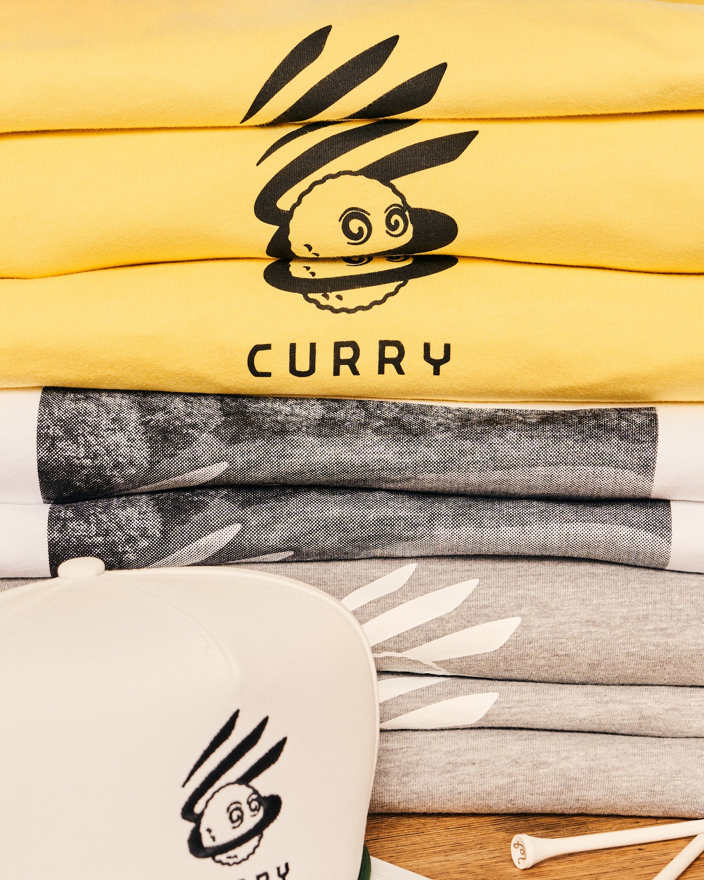 黃色 T 恤上的小人圖案與 CURRY 字樣,旁邊是灰色系與照片印花款|圖片來源:Malbon 官方 Instagram Curry Brand × Malbon 聯名黃色 T 恤與帽 T 疊放,印著 CURRY 小人圖案